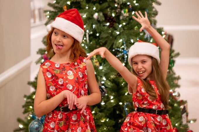 Two girls in red Santa-themed dresses and hats stand in front of a decorated Christmas tree.