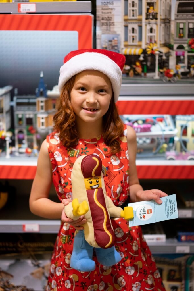 A smiling child in a festive red outfit and Santa hat holding a plush hot dog toy.