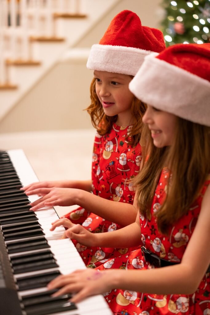 Children wearing Santa hats and red festive dresses playing a piano near a Christmas tree.