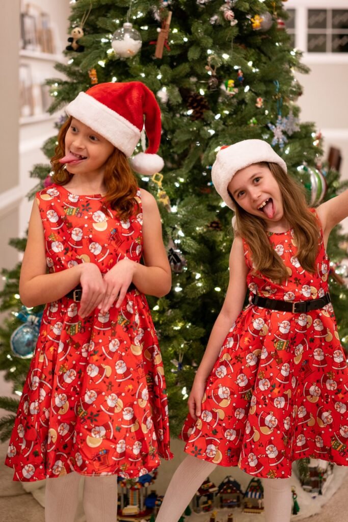 Two people modeling matching red floral dresses and Santa hats in front of a Christmas tree.