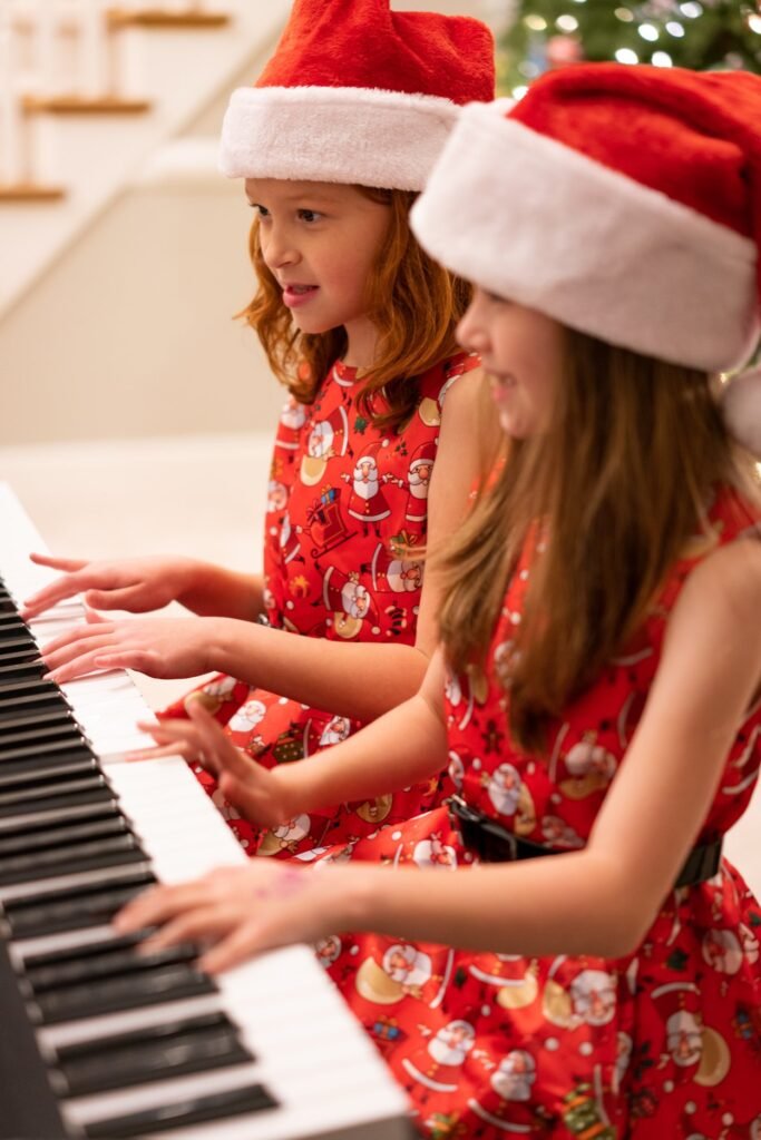 Three children in festive red dresses and Santa hats play the piano together. They are seated in a row in front of a decorated Christmas tree.