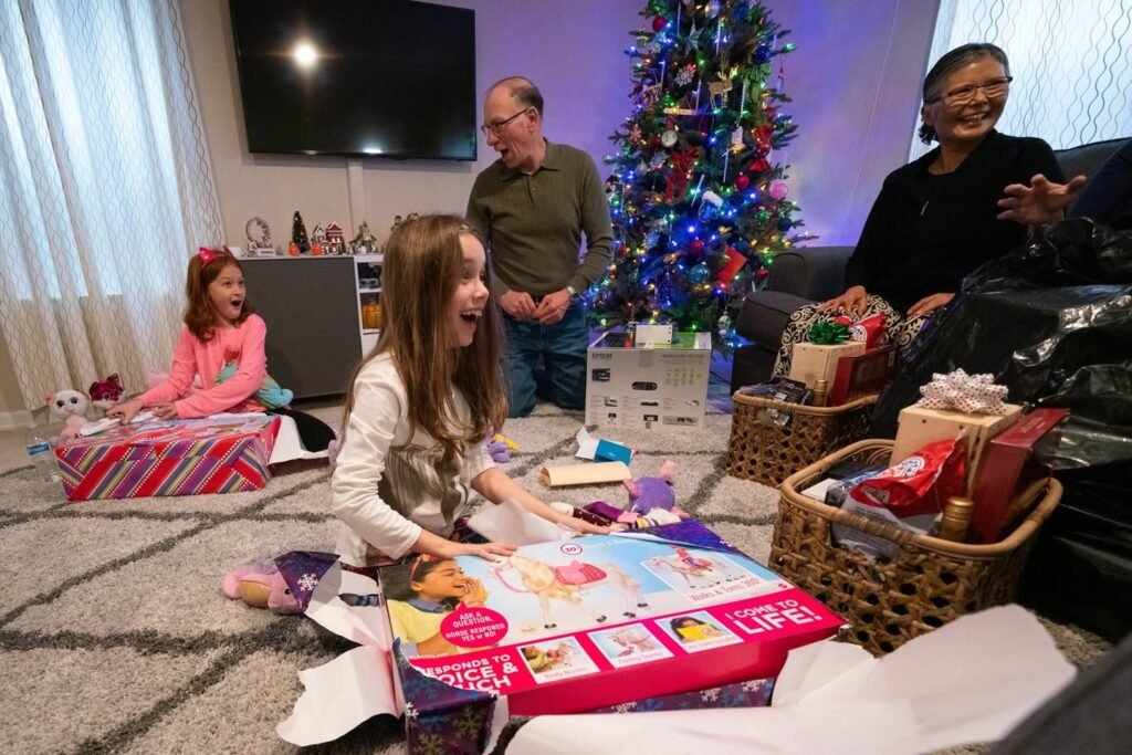 Family members enjoy Christmas morning gift-opening with a festive tree in the background. A young child opens a colorful gift on a carpeted floor.