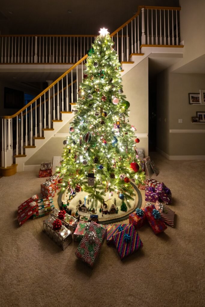 A tall, brightly lit Christmas tree decorated with ornaments and surrounded by colorful gift-wrapped presents indoors.