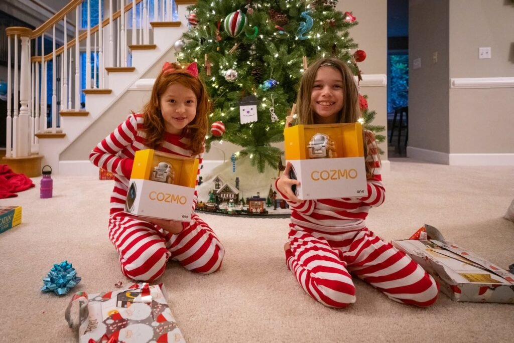 Two people in festive red and white striped pajamas sit on a carpet displaying yellow and white product boxes near a decorated Christmas tree. Background features a modern staircase with white balusters and wood accents.