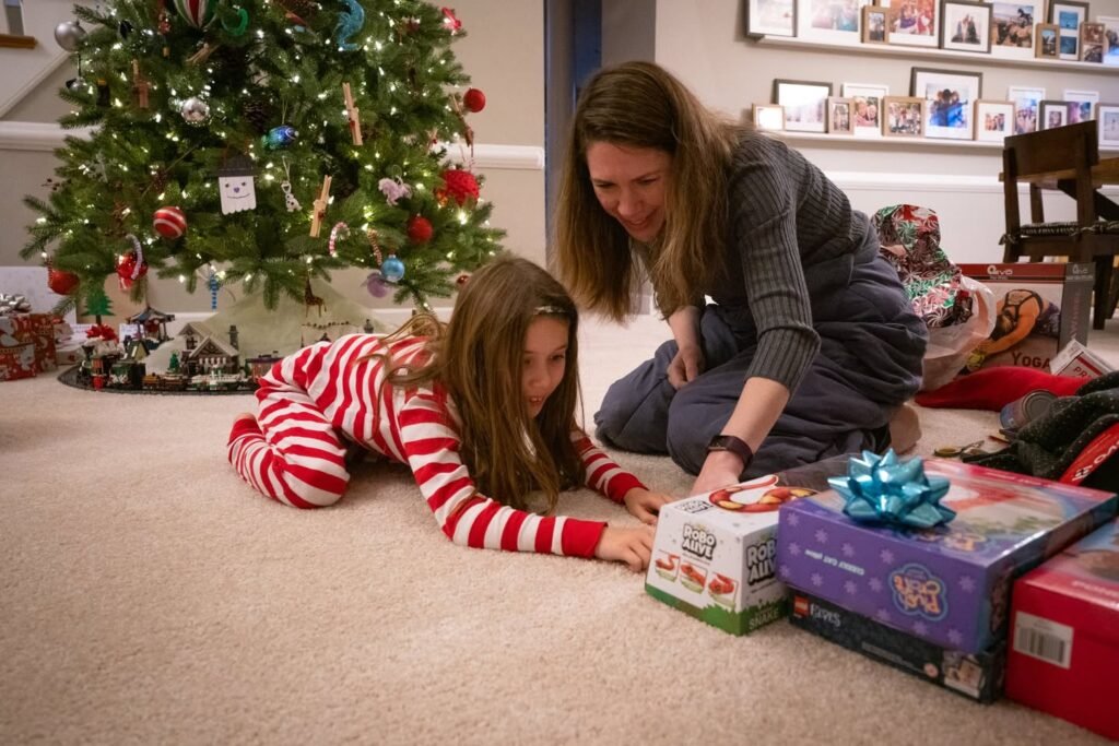 Two people opening gifts on a carpeted floor beside a decorated Christmas tree.