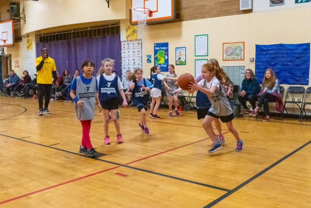 A group of young children playing basketball on an indoor court with a coach in a yellow shirt and spectators seated around. The gym features educational posters and a wooden floor.