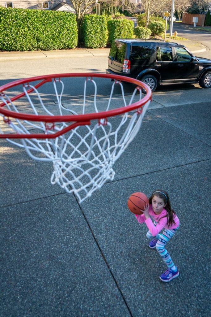 A child holds a basketball near a red rim on a sunny residential street.