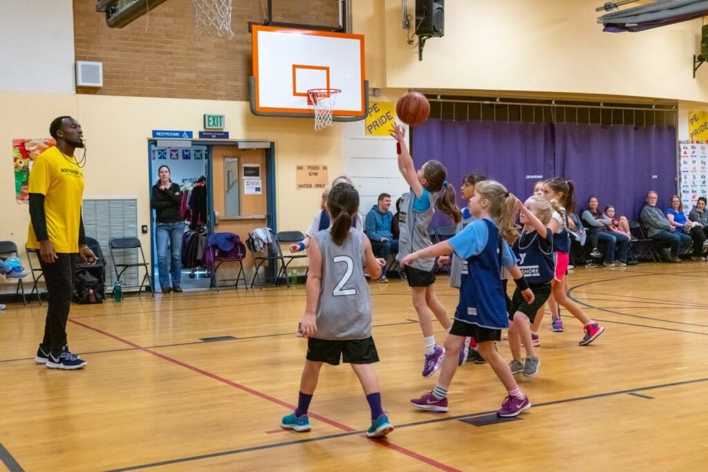 Young basketball players in a gymnasium with one player shooting the ball towards the hoop during a game with a referee and spectators. The scene captures active gameplay on a polished wooden court.