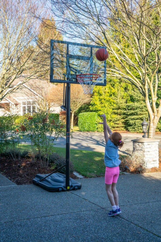 A child attempts to sink a basket on a backyard hoop, with trees and a home in the background.