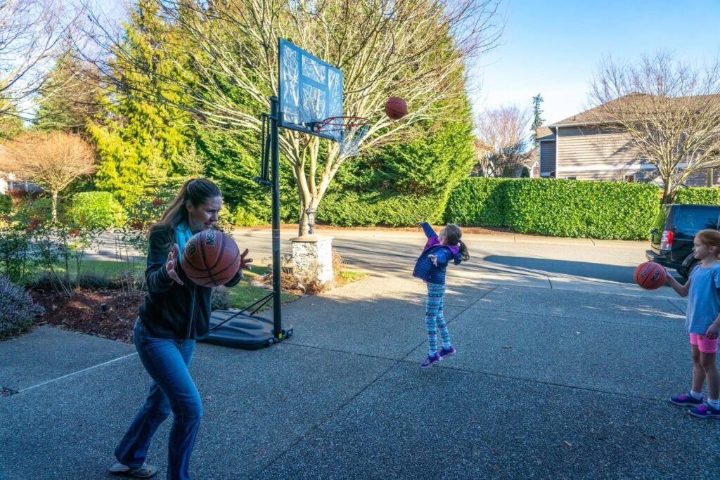 A family enjoys active playtime with basketballs on a residential driveway. The backyard features a basketball hoop, green hedges, and mature trees under a bright sky.