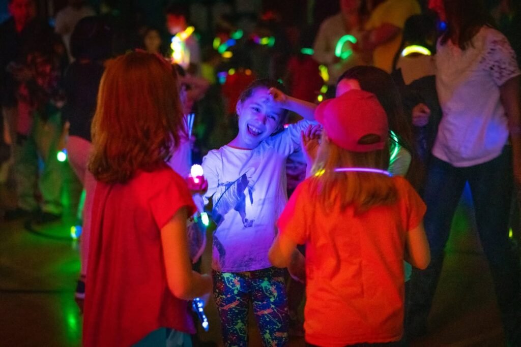 Kids wearing bright glow-in-the-dark bands and bracelets under colorful blacklight illumination at a nighttime outdoor event.