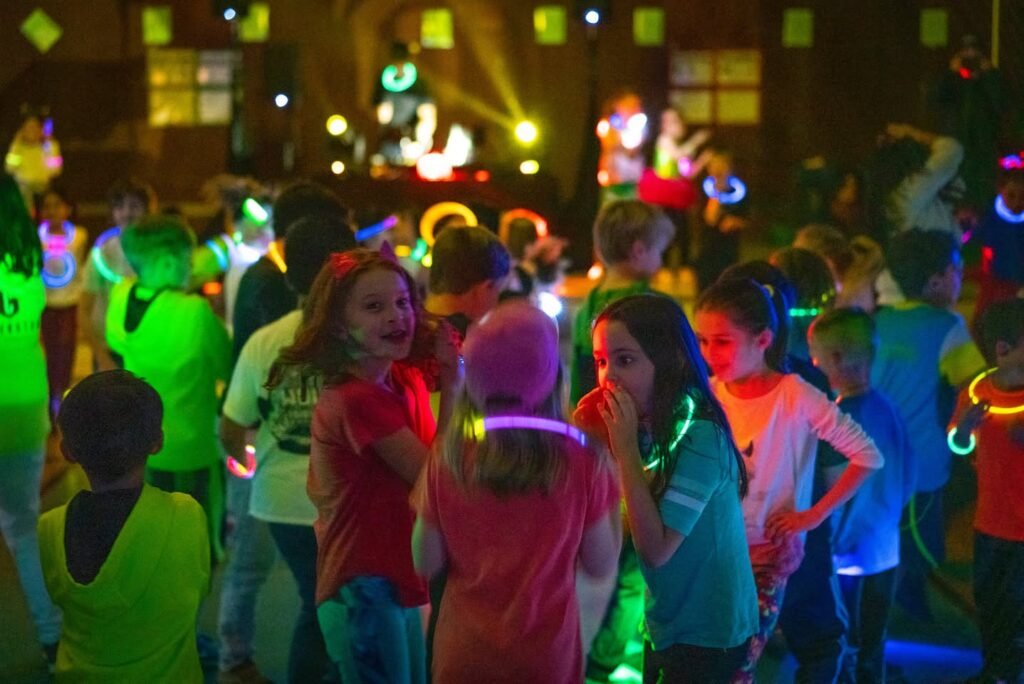 Children wearing glow sticks and night lights dance under blacklight effects during a lively indoor social gathering.