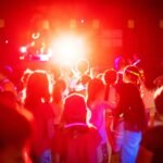An energetic audience at a concert, illuminated by intense red stage lights and a bright spotlight from the back.