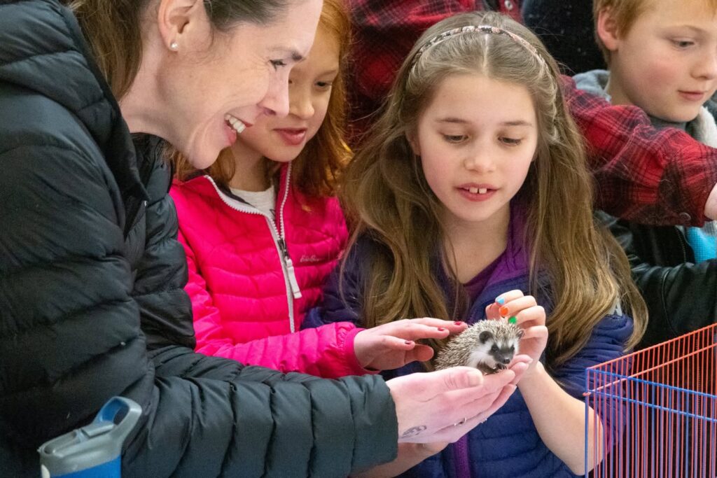 A group of children and adults gather around a small animal, possibly a ferret, held in someone's hands.