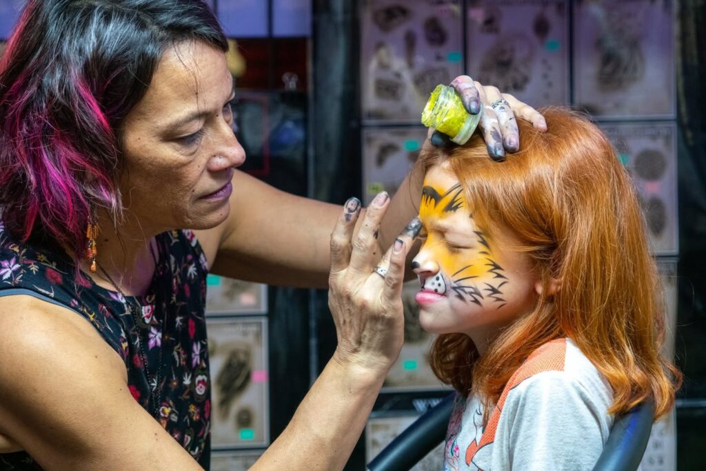 An artist applies tiger face paint to a young child with red hair. This photo captures a creative moment of fun for kids at a community gathering.