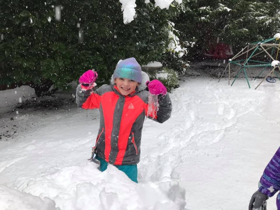 Person dressed in colorful winter gear playing in the snow. Snow-covered evergreen trees in the background of the yard.