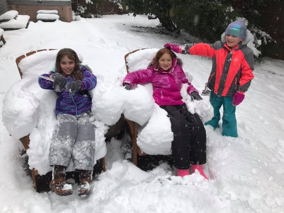 Children bundled up in winter jackets sitting on and standing near patio chairs completely covered in deep snow.
