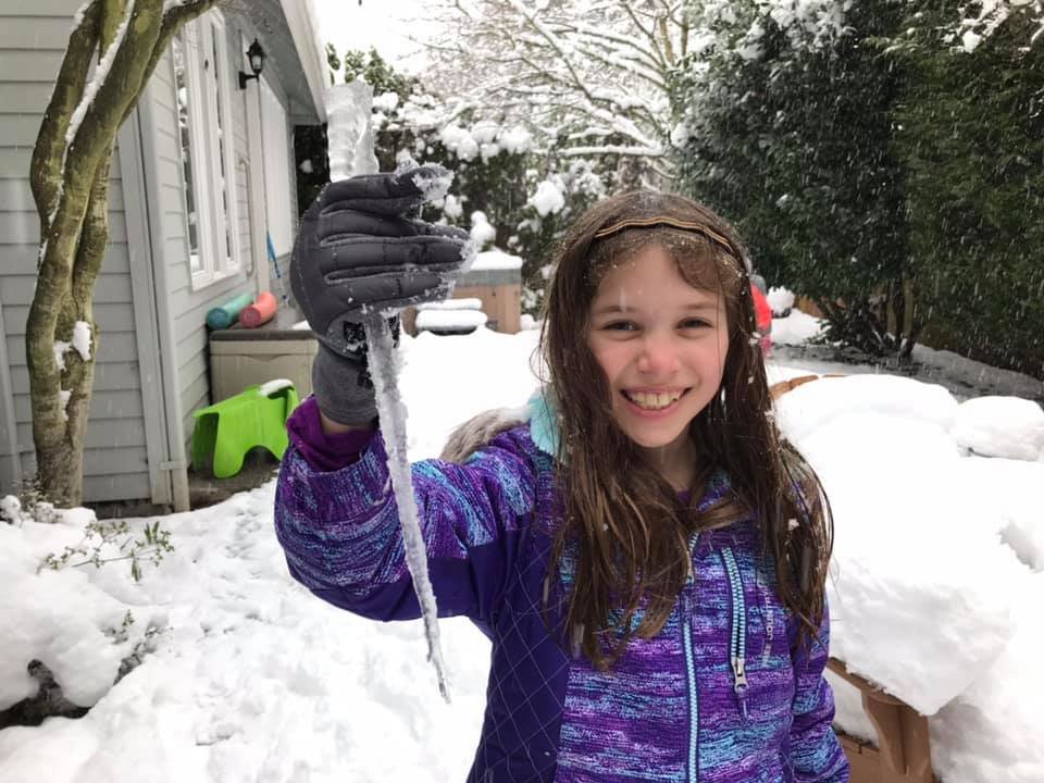 A young person with long hair proudly holds up a long, clear piece of ice in a snowy yard. The background shows a house and snow-covered trees.