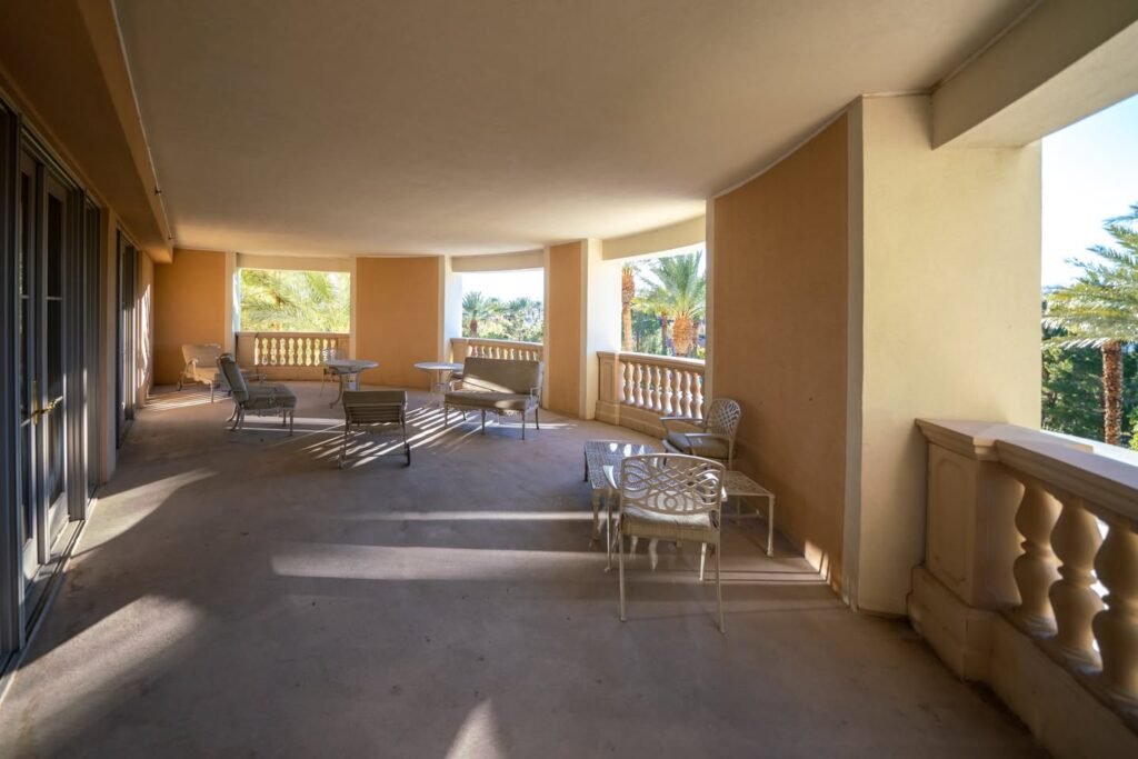 A sunny balcony with seating, a table, and a columned corner overlooking green foliage.