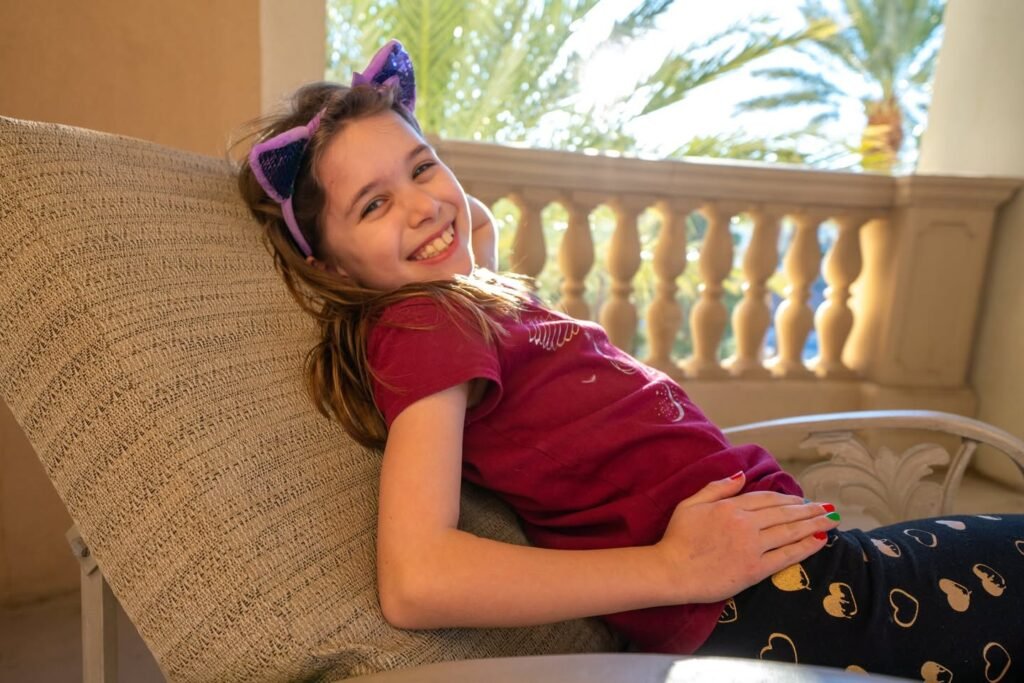 A child with sunglasses on their head smiles while resting in a beige outdoor chair with palm and baluster background
