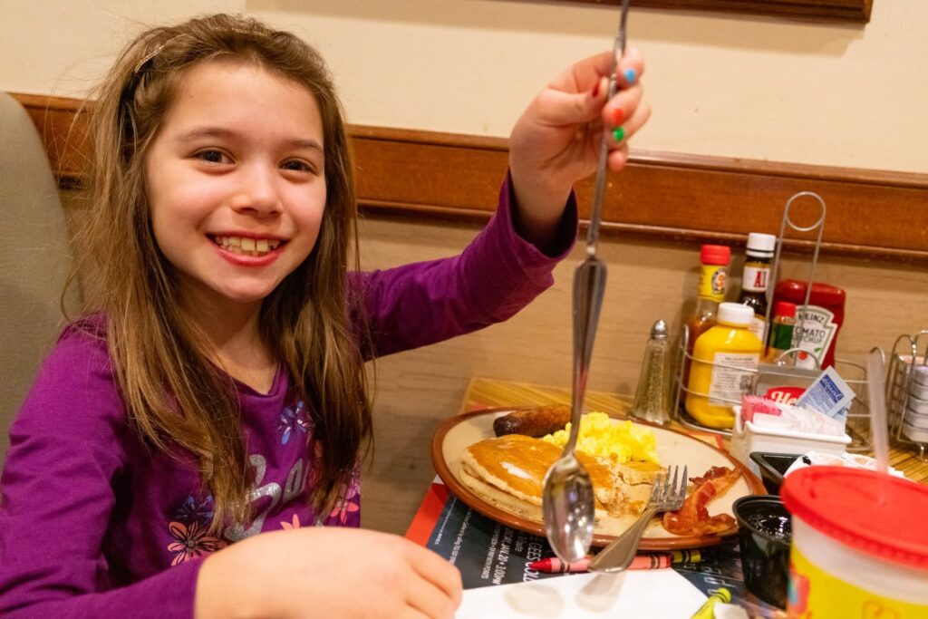 A smiling child in a purple shirt is seated at a diner table, holding up a fork with eggs and toast on the plate.