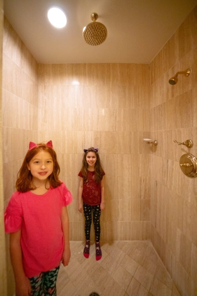 Kids wearing cat costume accessories in a large tiled shower room with brass fixtures.