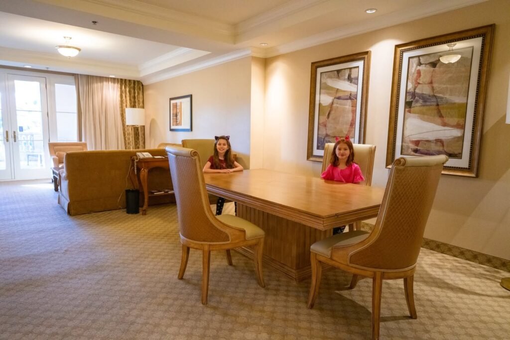 Spacious hotel suite with a wooden dining table and chairs, framed artwork on the walls, and two individuals seated at the table. Additional details include a couch and floor-to-ceiling windows.