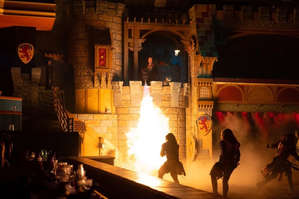 Actors in medieval costumes perform a pyrotechnic scene with a large fireball erupting on a stage designed to look like a castle.