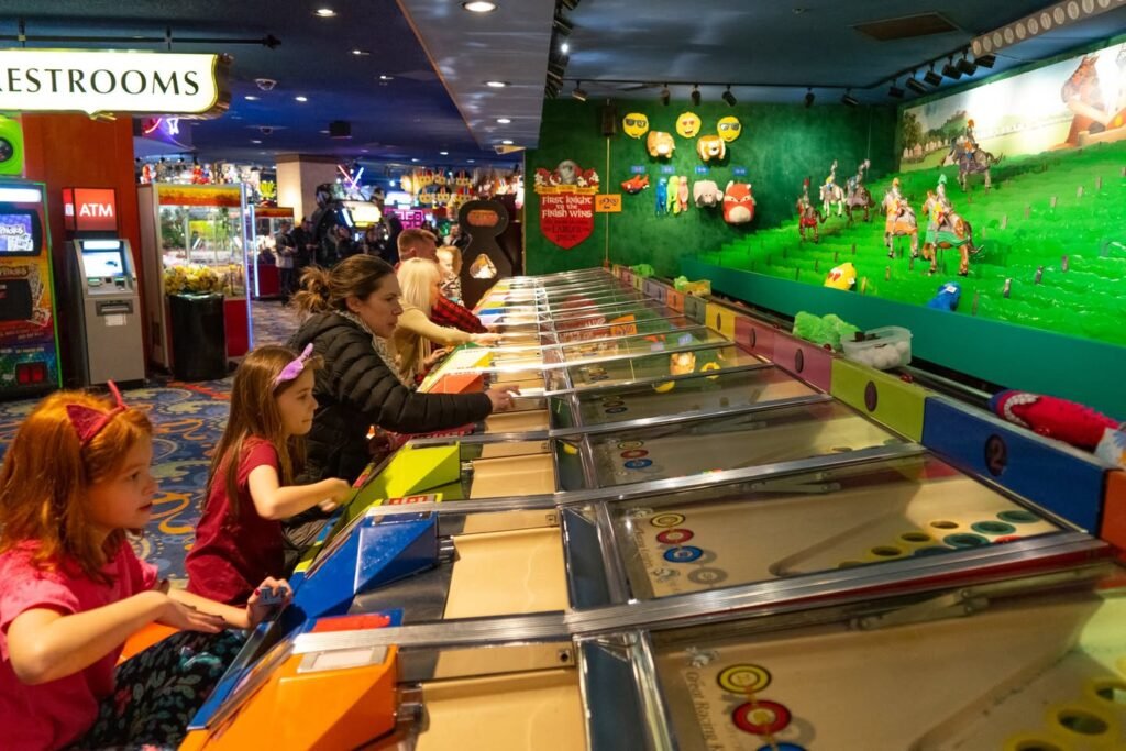 A row of colorful mini-golf lanes inside a Chuck E. Cheese restaurant with players lining the course. A 'Restrooms' sign and various arcade machines are visible in the background.