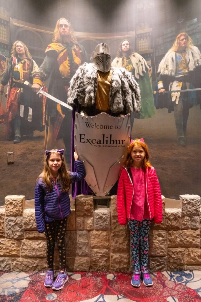 Two children stand in front of a misty medieval 'Welcome to Excalibur' display. Figures in knight and armored costumes are visible in the background.