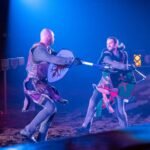 Actors performing with swords and shields on a blue-lit stage.