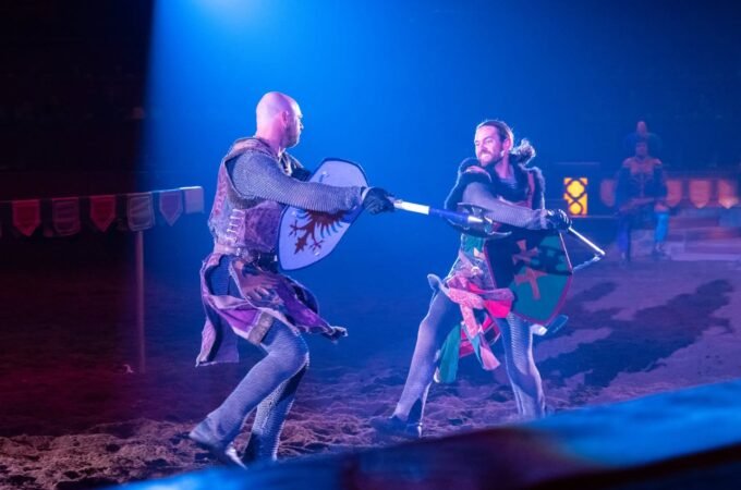 Actors performing with swords and shields on a blue-lit stage.