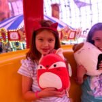 Container Park Children holding plush toys at a carnival ride, with carousel lights in the background.