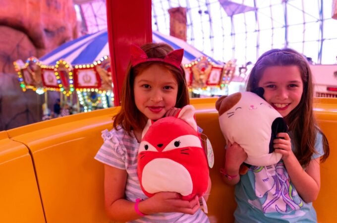 Children holding plush toys at a carnival ride, with carousel lights in the background.