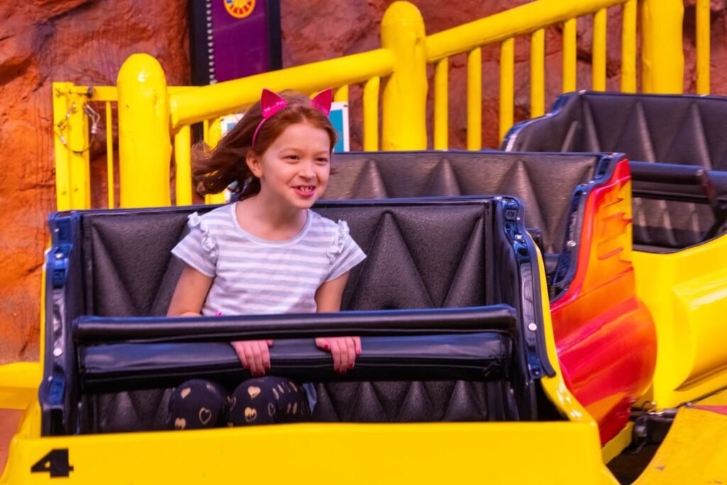 Young girl enjoying a ride on a yellow and black park coaster, with visible safety bar and rock-themed backdrop.