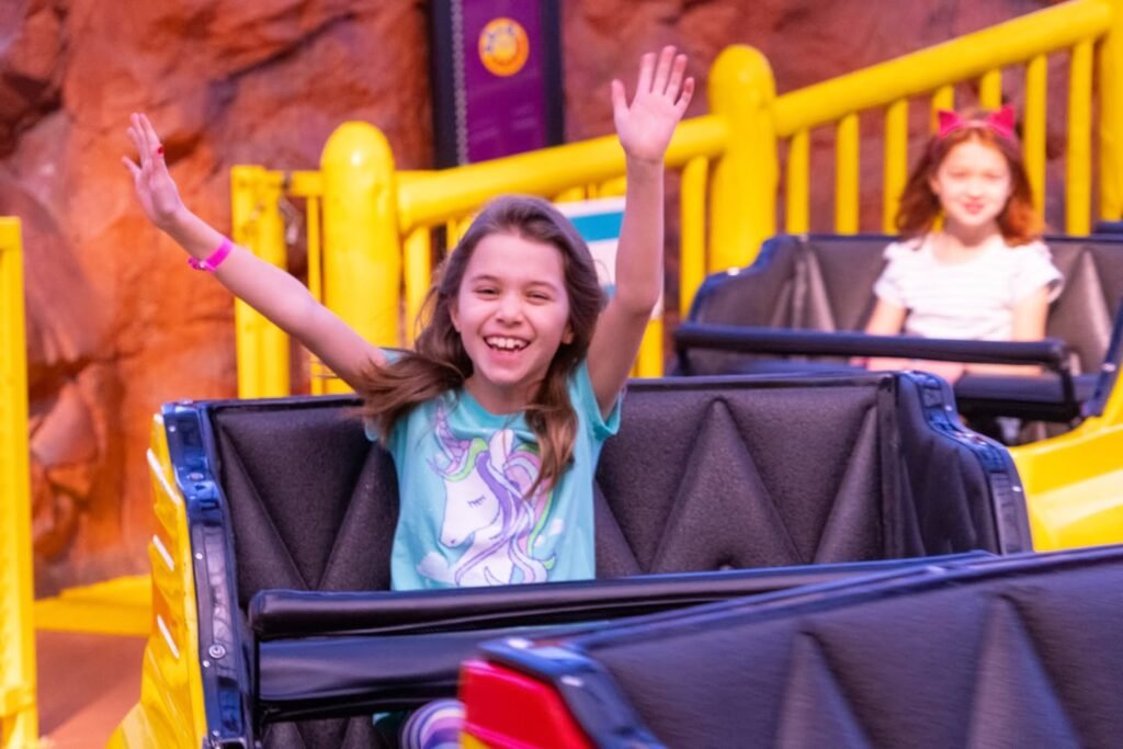 Two young girls riding a roller coaster with arms raised, enjoying the thrill at a theme park.