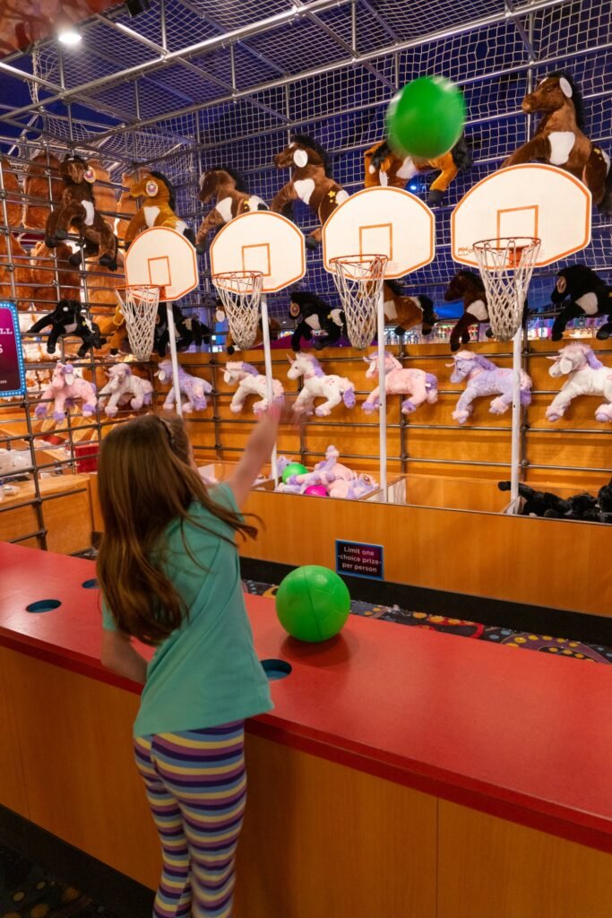 A person playing an arcade basketball game with plush toy prizes in an indoor entertainment facility.