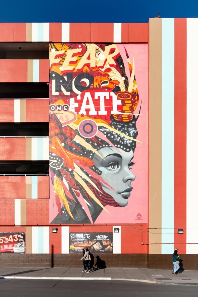 A close-up view of a colorful street mural featuring a woman with an elaborate headdress and text that reads 'FEAR NO FAITH.' The artwork is displayed on a building with bold red, orange, and white stripes.