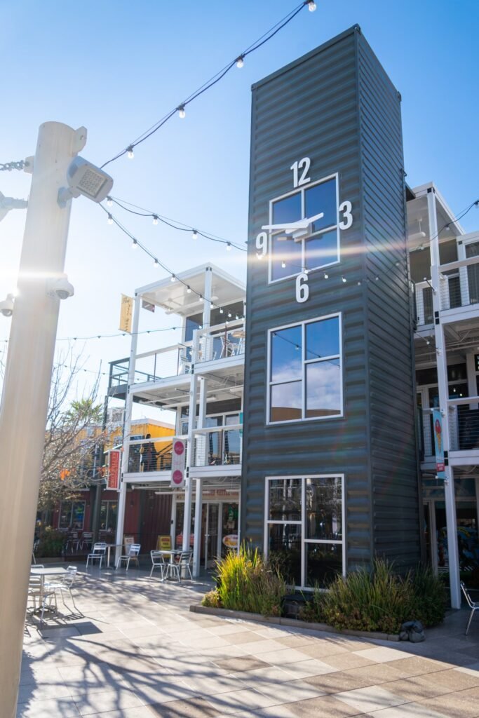 A tall grey clock tower stands between multi-story buildings under a clear blue sky with string lights overhead.