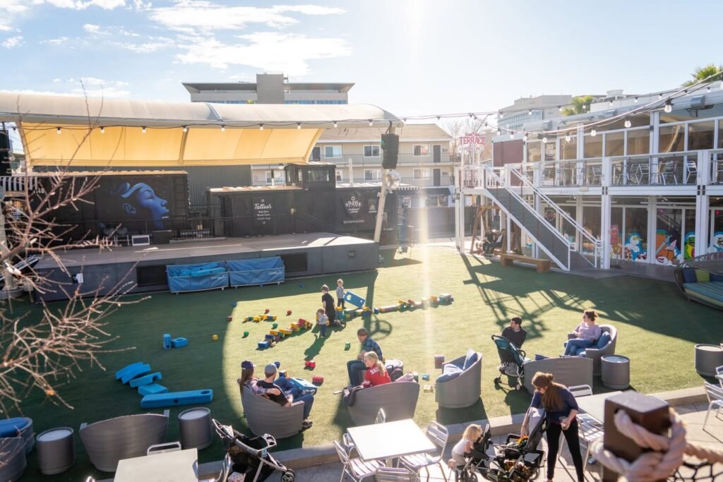 A lively rooftop space with a grassy area, colorful play structures, and outdoor seating. People are relaxed on the lawn, with the city skyline visible in the distance.