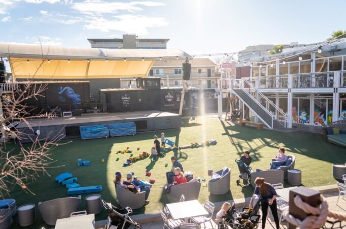 A lively rooftop space with a grassy area, colorful play structures, and outdoor seating. People are relaxed on the lawn, with the city skyline visible in the distance.