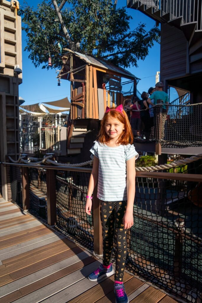 A person in patterned leggings stands on a wooden boardwalk near theme park structures and a large tree against a blue sky