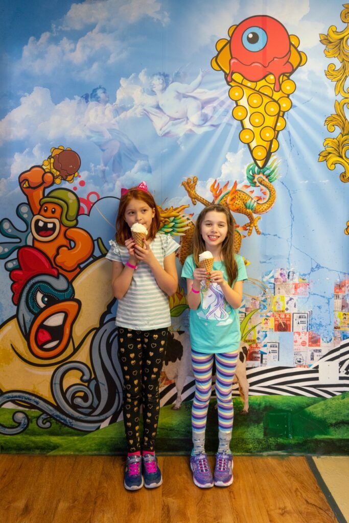 Two children enjoy ice cream in front of a colorful mural featuring a giant ice cream cone and cartoon characters.