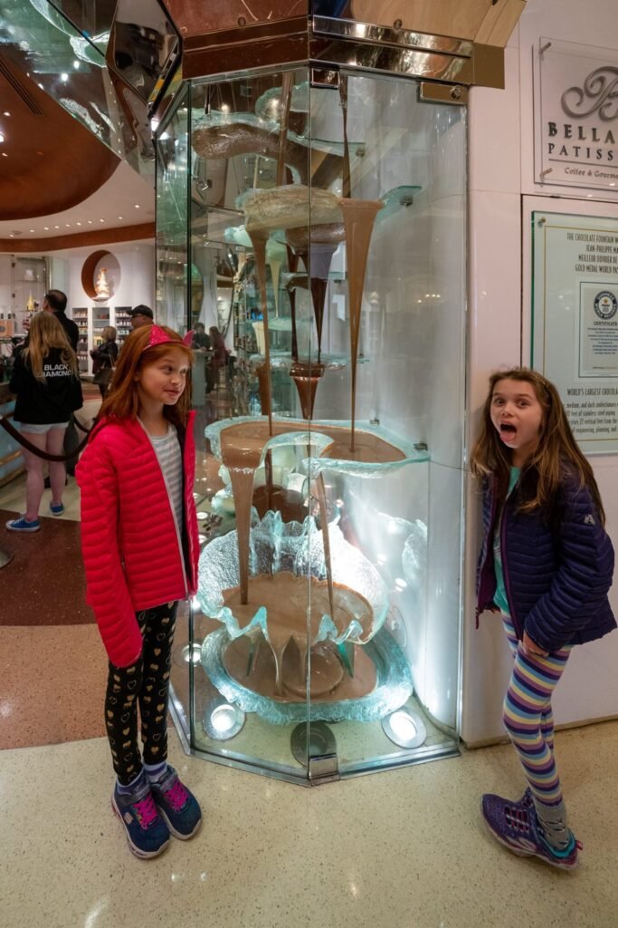 Two children explore a large glass-enclosed hot chocolate fountain with one showing surprise. The indoor setting features warm lighting and a Bella Fatini sign in the background.