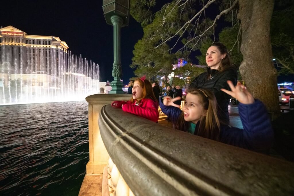 Visitors view the illuminated Bellagio fountains at night from a waterfront railing.