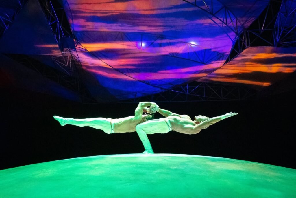 A performer in a dynamic pose under a vividly colored stage backdrop. The lighting creates a surreal and dramatic atmosphere.