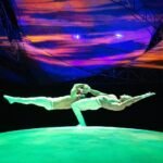 A performer in a dynamic pose under a vividly colored stage backdrop. The lighting creates a surreal and dramatic atmosphere.