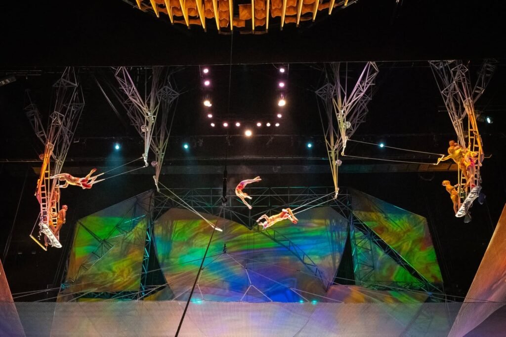 Acrobats performing high-flying circus routines on a colorful stage, with elaborate lighting and intricate aerial rigging.
