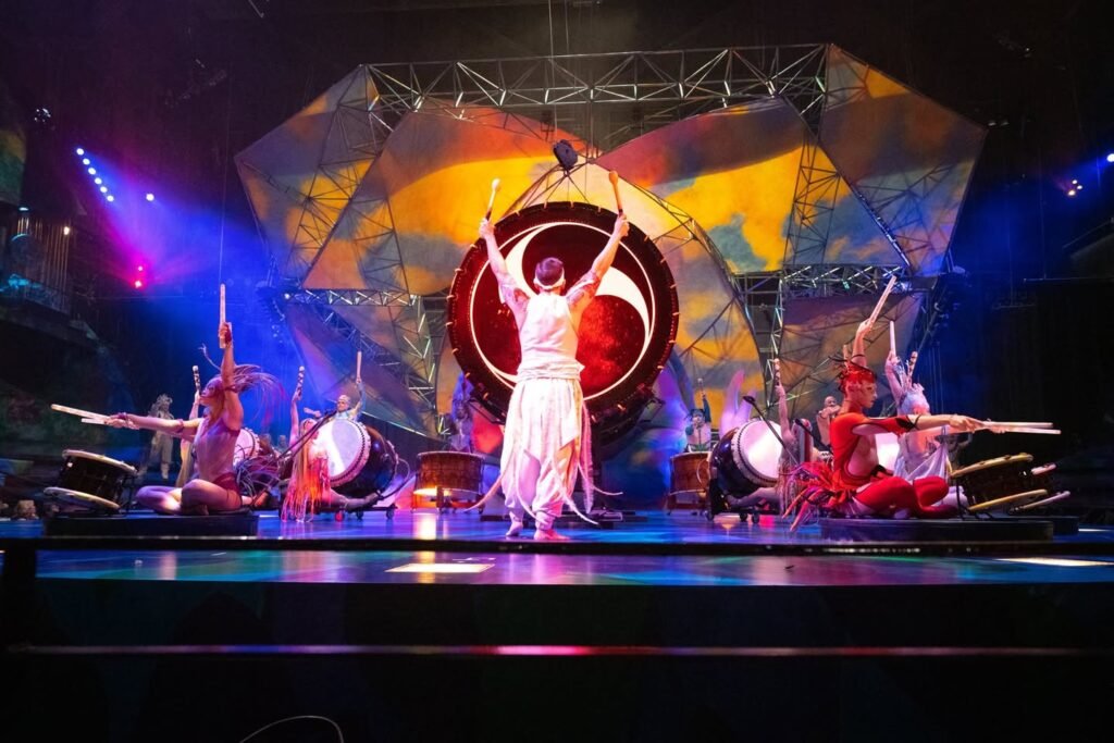 A performer with a drum stick raises their arms on stage surrounded by a percussion ensemble during a live concert. Colorful lighting and artistic backdrops enhance the venue atmosphere.