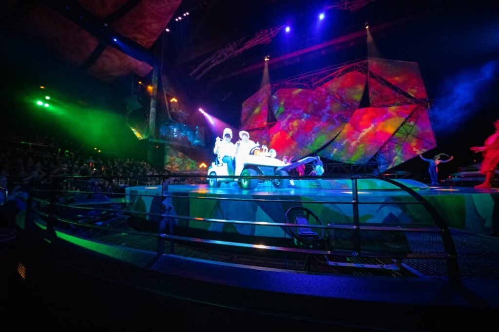 A colorful concert stage with vibrant lighting and a ship-themed backdrop. Dynamic green and blue lights illuminate the scene.
